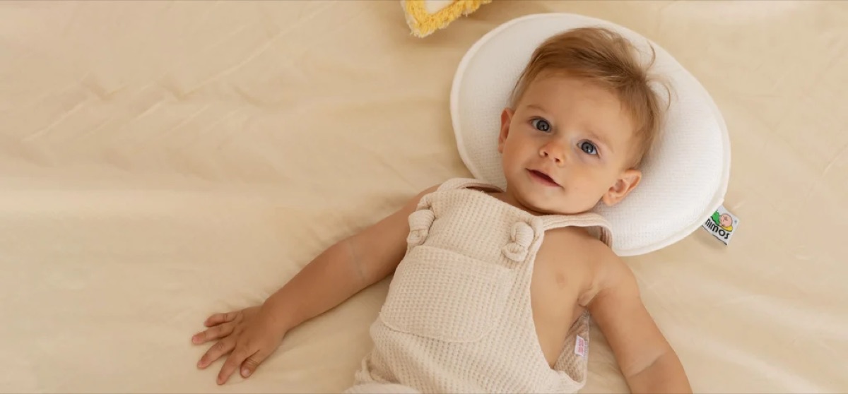 An infant lying on their back with the head resting in the central recess of a white oval head-shaping pillow