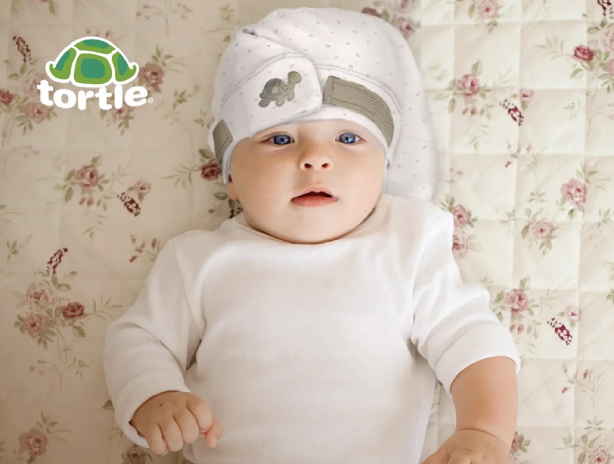 An infant lying on their back wearing a soft beanie with a side strap and a contoured roll positioned along one side of the head