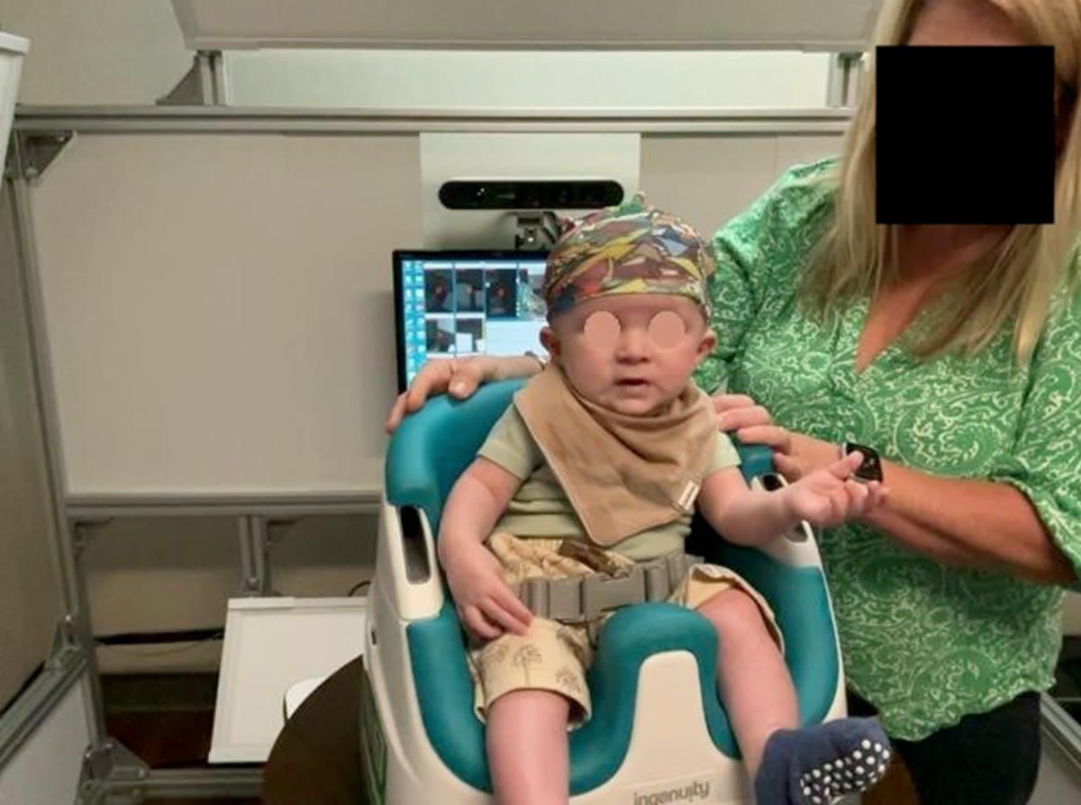 A SoftSpot3D scan session: an infant in the patterned cap sits in the baby seat on the turntable, facing the depth sensor, while an operator steadies the seat. Faces are masked.
